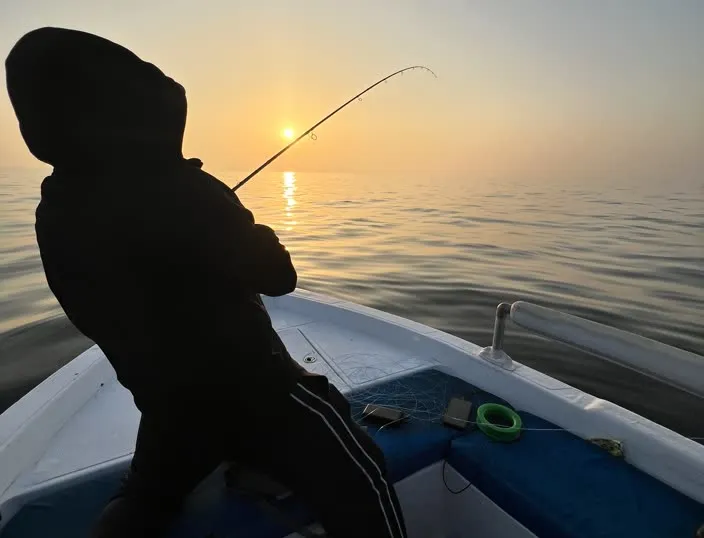 A silhouetted person fishing from a small boat at sunrise.